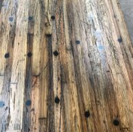 A close-up of a wooden table with a rustic appearance. The surface has a weathered texture with visible wood grain and scattered dark circular markings. The color varies from light to dark brown tones.