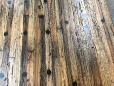 A close-up of a wooden table surface featuring a rustic design with visible grain patterns and scattered black circular markings. The wood appears weathered and textured, contributing to its vintage look.