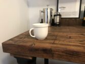 A white coffee mug sits on a rustic wooden table. Behind it, theres a French press, a coffee grinder, and a framed definition of coffee on the wall. The setting is minimalistic with a cozy, homey feel.