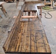 A long, L-shaped wooden bench under construction in a workshop. The surface appears worn and marked with visible scratches and cuts. In the background, there are tools, lumber, and coiled cables.