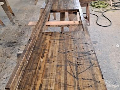 A long, L-shaped wooden bench under construction in a workshop. The surface appears worn and marked with visible scratches and cuts. In the background, there are tools, lumber, and coiled cables.