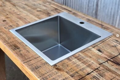A square metal sink is installed in a rustic wooden countertop. The background features a corrugated metal wall, adding an industrial feel to the setting.