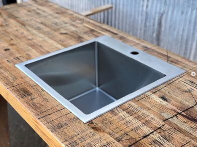 A square metal sink is installed in a rustic wooden countertop. The background features a corrugated metal wall, adding an industrial feel to the setting.