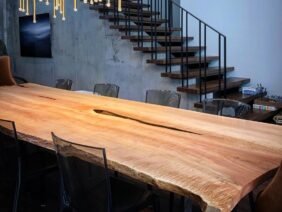 A modern dining room features a long wooden table with a natural edge, surrounded by transparent chairs. Above, numerous slender pendant lights hang in an artistic cluster. A staircase with black railings ascends in the background.