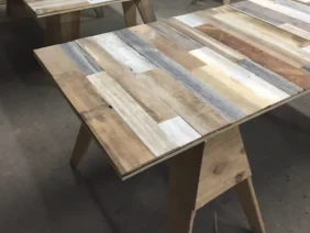 Wooden tables with plank-style surfaces and sawhorse legs are arranged in a workshop. Each table features a variety of wood tones and textures, creating a rustic and handcrafted appearance.