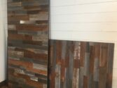 Two weathered wooden panels with a mix of brown, gray, and blue tones lean against a white shiplap wall on a wooden floor.