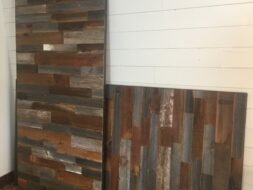 Two weathered wooden panels with a mix of brown, gray, and blue tones lean against a white shiplap wall on a wooden floor.