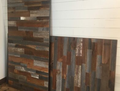 Two weathered wooden panels with a mix of brown, gray, and blue tones lean against a white shiplap wall on a wooden floor.