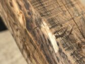 Close-up of a rough, worn wooden beam with visible grain patterns and knots; the surface shows signs of weathering and aging. The background is blurred, emphasizing the texture and detail of the wood.