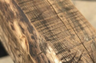 Close-up of a rough, worn wooden beam with visible grain patterns and knots; the surface shows signs of weathering and aging. The background is blurred, emphasizing the texture and detail of the wood.