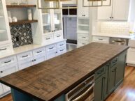 A modern kitchen with white cabinets, a wooden floor, and a central island featuring a dark wood countertop. There are built-in ovens in the island, and decorative pendant lights hang above.
