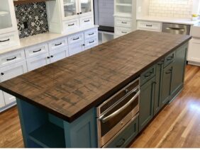 A modern kitchen with white cabinets, a wooden floor, and a central island featuring a dark wood countertop. There are built-in ovens in the island, and decorative pendant lights hang above.