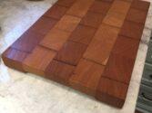 A wooden cutting board with a checkered pattern of alternating light and dark wood grains sits on a marble countertop. The board features square blocks, emphasizing the natural wood textures.