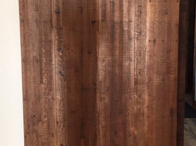 A large sliding barn door made of rustic wooden planks with a visible wood grain texture. It hangs from a black metal track and partially covers a wooden floor. The door has a warm, natural finish.
