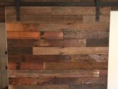 A wooden sliding barn door with a mix of dark and light brown planks, mounted on a black metal rail system, set against a white wall. The wood grain and texture are visible, giving a rustic appearance.