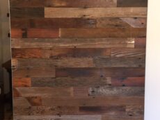 A wooden sliding barn door with a mix of dark and light brown planks, mounted on a black metal rail system, set against a white wall. The wood grain and texture are visible, giving a rustic appearance.