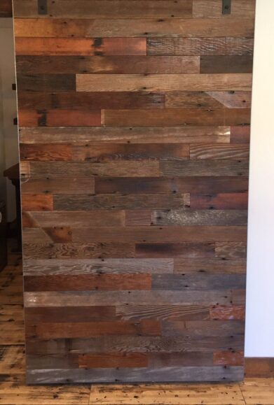 A wooden sliding barn door with a mix of dark and light brown planks, mounted on a black metal rail system, set against a white wall. The wood grain and texture are visible, giving a rustic appearance.