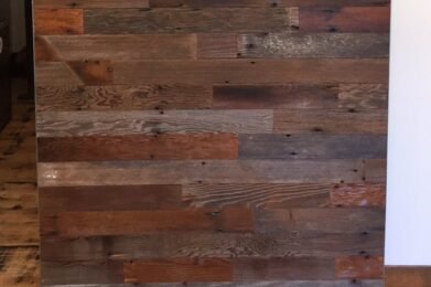 A wooden sliding barn door with a mix of dark and light brown planks, mounted on a black metal rail system, set against a white wall. The wood grain and texture are visible, giving a rustic appearance.