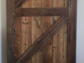 A rustic wooden barn door with a diagonal Z-brace design, made of weathered, vertically aligned wooden planks. It hangs on a black metal sliding track above a wooden floor. The door exudes a vintage, farmhouse aesthetic.