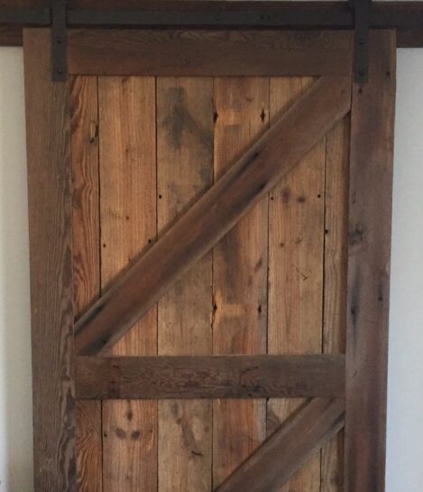 A rustic wooden barn door with a diagonal Z-brace design, made of weathered, vertically aligned wooden planks. It hangs on a black metal sliding track above a wooden floor. The door exudes a vintage, farmhouse aesthetic.