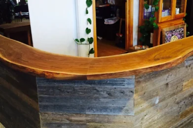 The Live Edge Tops reception desk curves around a corner, adorned with trailing green plants above. Behind it, shelves and cabinets accommodate various items in a warmly lit room that exudes a vintage charm. The floor is covered with small hexagonal tiles.