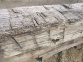Close-up of a weathered wooden beam with visible cracks and a small hole in the center. The surface appears rough and aged, resting on a sandy ground.