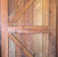 A rustic wooden barn door with vertical planks and a diagonal brace forming a Z pattern. The door displays a natural wood grain and warm brown tones, giving it a vintage, handcrafted appearance.