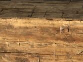 Close-up of a weathered wooden beam with a rough, textured surface. The wood displays signs of aging, including cracks and variations in color, creating an antique appearance.