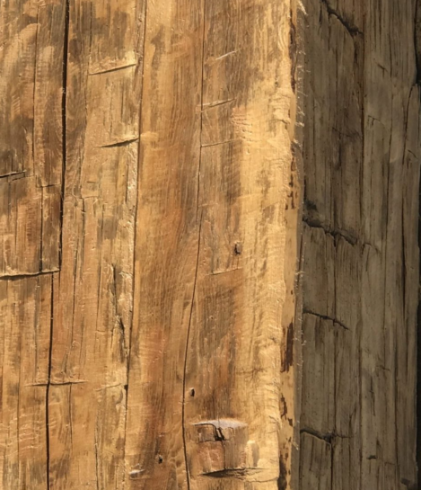 Close-up of a wooden beam showing natural textures, grooves, and some weathering. The woods warm brown tone and visible grain patterns suggest an aged, rustic appearance.