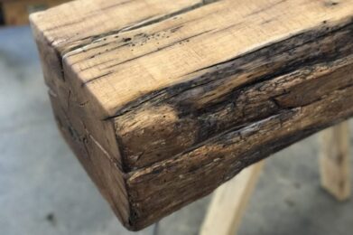 Close-up of a rustic wooden beam with visible grain and natural imperfections. The beam has a textured, weathered appearance, highlighting the rough edges and rich wood tones. It rests on a wooden surface.