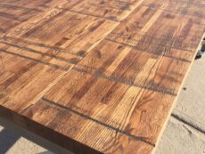 A close-up view of a tabletop with a rustic, wooden pattern. The surface features a variety of wood grain textures and a warm, natural finish. The table is set on a sunlit concrete surface.