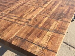 A close-up view of a tabletop with a rustic, wooden pattern. The surface features a variety of wood grain textures and a warm, natural finish. The table is set on a sunlit concrete surface.