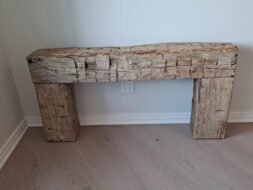 Rough-hewn wooden mantelpiece with a natural, rustic look, placed against a plain white wall on light wood flooring. An electrical outlet is visible on the wall behind it.