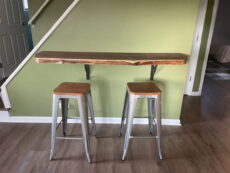 A minimalist bar setup with a wooden live-edge countertop mounted on a light green wall. Two metal bar stools with wooden seats are placed in front. The area has wooden flooring, and stairs are partially visible on the left.