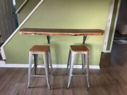 A minimalist bar setup with a wooden live-edge countertop mounted on a light green wall. Two metal bar stools with wooden seats are placed in front. The area has wooden flooring, and stairs are partially visible on the left.