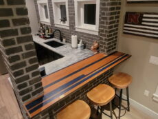 Home bar area with a brick wall, marble countertop, and three wooden stools. The bar top features an inlaid design with wood and dark stripes. Shelves with bottles and decor are visible behind the counter. Three small windows are set in the brick wall.