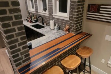 Home bar area with a brick wall, marble countertop, and three wooden stools. The bar top features an inlaid design with wood and dark stripes. Shelves with bottles and decor are visible behind the counter. Three small windows are set in the brick wall.