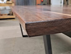 Close-up of a wooden table edge with a sleek, polished surface. The modern design features a smooth, angled corner and rests on a sturdy, minimalist metal leg. The background is slightly blurred.