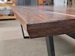 Close-up of a wooden table edge with a sleek, polished surface. The modern design features a smooth, angled corner and rests on a sturdy, minimalist metal leg. The background is slightly blurred.