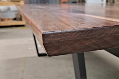 Close-up of a wooden table edge with a sleek, polished surface. The modern design features a smooth, angled corner and rests on a sturdy, minimalist metal leg. The background is slightly blurred.