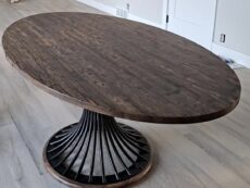 A large oval wooden dining table with a dark, polished surface and a unique pedestal base made of curved metal bars, set on a light wood floor in a room with beige walls and a white door.