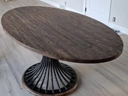 A large oval wooden dining table with a dark, polished surface and a unique pedestal base made of curved metal bars, set on a light wood floor in a room with beige walls and a white door.