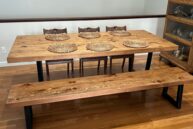 A wooden dining table set with six intricately designed wicker placemats, accompanied by a matching bench and three wooden chairs, all set on a polished hardwood floor. A shelf with glassware is visible on the right.