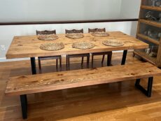 A wooden dining table set with six intricately designed wicker placemats, accompanied by a matching bench and three wooden chairs, all set on a polished hardwood floor. A shelf with glassware is visible on the right.