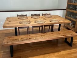A wooden dining table set with six intricately designed wicker placemats, accompanied by a matching bench and three wooden chairs, all set on a polished hardwood floor. A shelf with glassware is visible on the right.