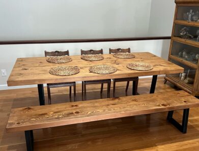 A wooden dining table set with six intricately designed wicker placemats, accompanied by a matching bench and three wooden chairs, all set on a polished hardwood floor. A shelf with glassware is visible on the right.