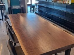 A large, rustic wooden table with a natural edge, surrounded by dark high-back chairs in a modern dining area. Shelves with bottles line the wall, and large windows in the background let in natural light.