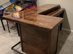 A polished wooden bar with a corner design stands on a carpeted floor, adjacent to vintage pinball machines with colorful designs. A metal footrest is attached to the bottom of the bar.