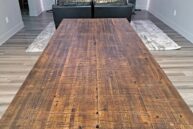 A long wooden dining table with a rustic finish in a modern living room. The room features wood flooring, two large speakers, a TV on the wall, and a leather sofa set on a textured rug.