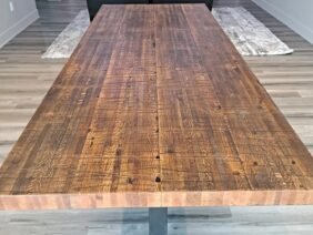 A long wooden dining table with a rustic finish in a modern living room. The room features wood flooring, two large speakers, a TV on the wall, and a leather sofa set on a textured rug.